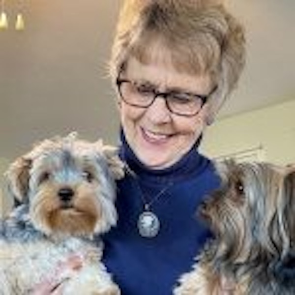 A smiling Nancy Weaver wearing eyeglasses and a blue turtleneck while holding two small dogs.